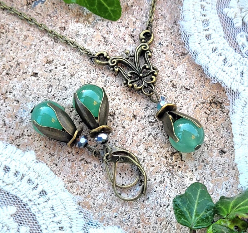 Pode incluir: Um colar com pingente de vidro verde e brincos combinando com um design de filigrana de bronze. O pingente apresenta um design de borboleta.