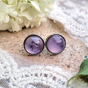 Può includere: Un paio di orecchini a bottone in argento con un cabochon di vetro viola chiaro in ogni orecchino. I cabochon hanno un aspetto leggermente nuvoloso.