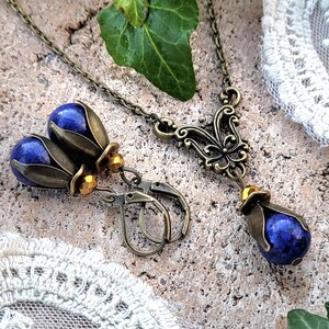 Pode incluir: Um par de brincos e um colar com pedras de l&aacute;pis-laz&uacute;li azuis engastadas em desenhos florais de bronze ornamentados. Os brincos t&ecirc;m um design de gota com um pequeno acento de contas douradas. O colar tem um pingente com um design de borboleta.