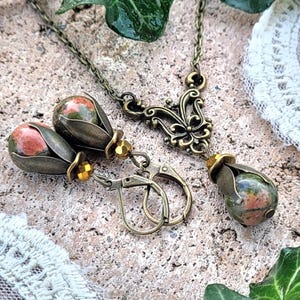Pode incluir: Um conjunto de brincos e um colar com uma pedra em forma de l&aacute;grima com padr&otilde;es verdes, rosa e marrons. As joias s&atilde;o feitas de metal de cor bronze e t&ecirc;m um design floral.