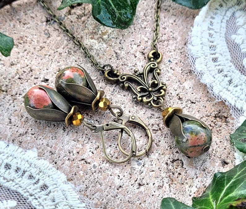 Pode incluir: Um conjunto de brincos e um colar com uma pedra em forma de l&aacute;grima com padr&otilde;es verdes, rosa e marrons. As joias s&atilde;o feitas de metal de cor bronze e t&ecirc;m um design floral.