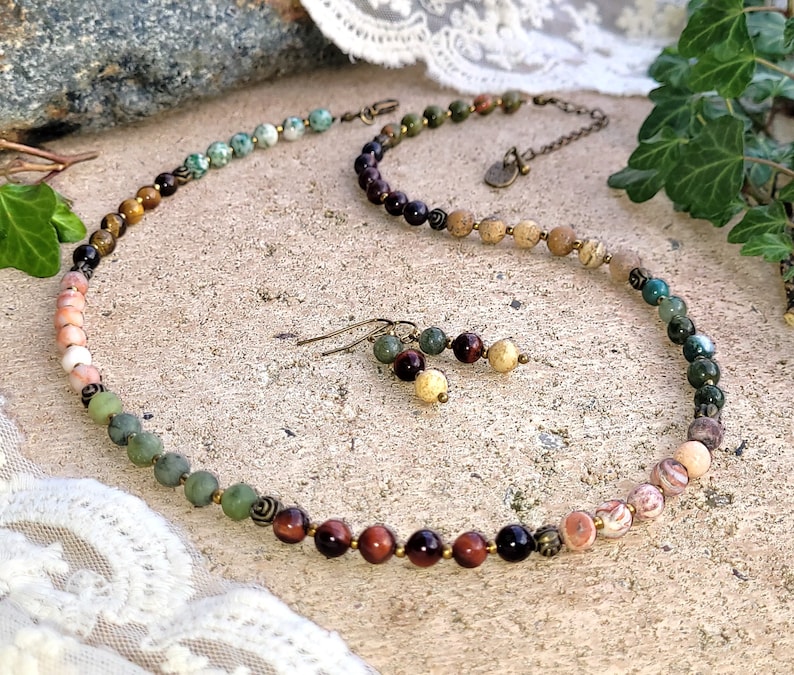 May include: A multi-colored gemstone necklace with matching earrings. The necklace features a variety of stones, including tiger's eye, red jasper, and green aventurine. The earrings are made with the same stones as the necklace.