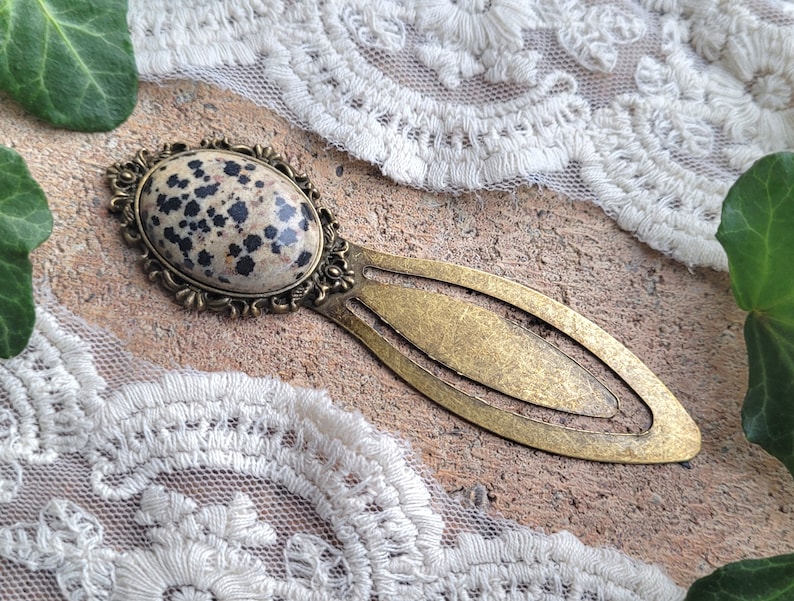 Op de afbeelding: Een messing bladwijzer met een inleg van dalmatijnse jaspis. De bladwijzer heeft de vorm van een lange ovaal met een decoratief ontwerp rond de steen.