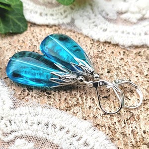 Pode incluir: Um par de brincos em forma de lágrima com uma configuração de metal prateado. Os brincos apresentam uma grande pedra de vidro em forma de lágrima de cor azul brilhante.