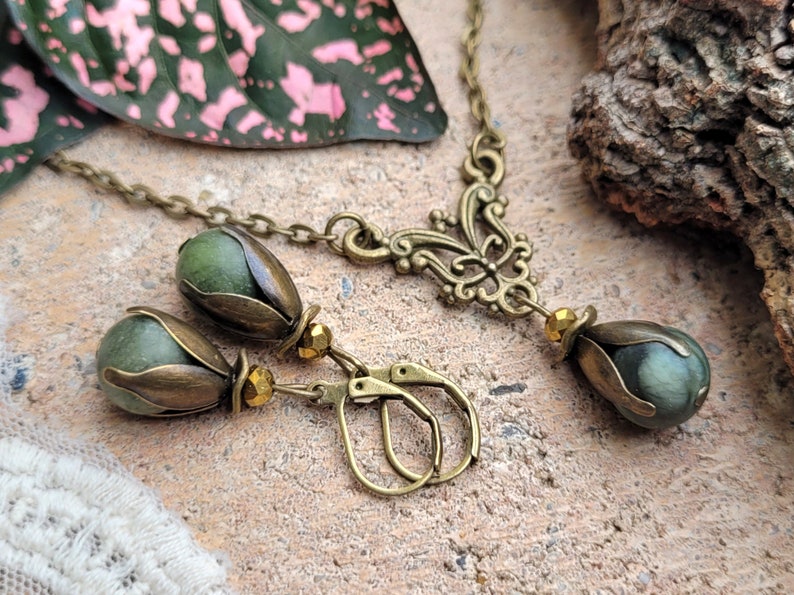 Pode incluir: Um conjunto de colar e brincos verde e bronze com um design floral. O colar tem um pingente com uma pedra verde e os brincos t&ecirc;m pedras verdes combinando.