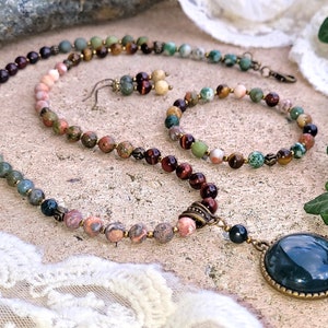 May include: A multi-colored gemstone necklace with a large green agate pendant. The necklace is made with various stones, including tiger's eye, jasper, and aventurine. The necklace is paired with a matching bracelet and earrings.