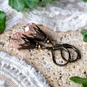 Könnte beinhalten: Ein Paar rosa Kristallohrringe mit bronzenen Akzenten und einem zarten, verzierten Design. Die Ohrringe haben einen tropfenförmigen Kristall mit einer facettierten Oberfläche und einen bronzefarbenen Draht, der um den Kristall gewickelt ist. Der bronzefarbene Draht ist mit kleinen, dekorativen Perlen verziert. Die Ohrringe sind an bronzefarbenen Bügelverschlüssen befestigt.