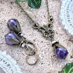 Pode incluir: Um par de brincos e um colar com pedras preciosas de ametista engastadas em desenhos florais de bronze antigo. Os brincos t&ecirc;m a forma de uma l&aacute;grima e pendem de ganchos de bronze. O colar tem um design delicado e ornamentado com um pingente em forma de l&aacute;grima de ametista.