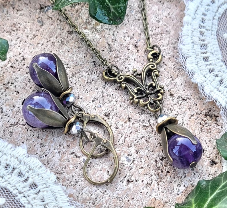 Pode incluir: Um par de brincos e um colar com pedras preciosas de ametista engastadas em desenhos florais de bronze antigo. Os brincos t&ecirc;m a forma de uma l&aacute;grima e pendem de ganchos de bronze. O colar tem um design delicado e ornamentado com um pingente em forma de l&aacute;grima de ametista.