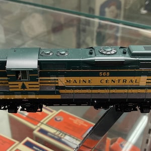 Puede incluir: Locomotora de tren modelo en verde oscuro y amarillo, con las palabras "MAINE CENTRAL" y el número "568" impresos en el lateral. El modelo tiene detalles y se exhibe en una vitrina.