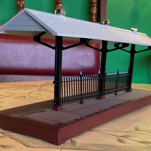 Puede incluir: Una plataforma de estación de tren modelo con un techo gris y soportes negros. La plataforma tiene una valla de metal negro y una base marrón.