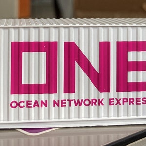 Puede incluir: Un contenedor de envío blanco con la palabra "ONE" en letras grandes de color rosa y el texto "OCEAN NETWORK EXPRESS" debajo. El contenedor tiene una señal de advertencia amarilla en el lateral.