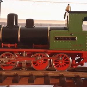 Puede incluir: Un tren de juguete negro y verde con ruedas rojas y ribetes dorados. El tren tiene un vagón de cola y las palabras "MAX Lok 01" en el lateral.