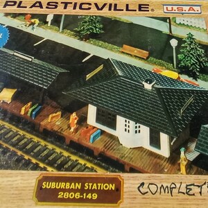Puede incluir: Una caja para un tren modelo de calibre HO con una estación suburbana con techo negro y paredes blancas. La caja está etiquetada como "Plasticville" y "The Original Plastic Village". La caja también dice "Snap Fit, No Glue" y "Complete".