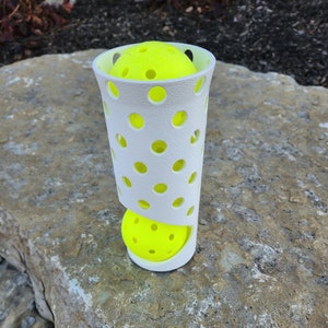 May include: A white plastic ball holder with a cylindrical shape and a pattern of holes. Two bright yellow balls are inside the holder.