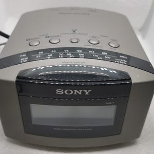 May include: A grey Sony digital clock radio with a black display and buttons for alarm and radio functions. The clock radio has a digital display and a radio frequency scale. The text "SONY" is visible on the front.