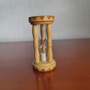 May include: A wooden hourglass with a light brown finish. The hourglass has a decorative design around the top and bottom. The glass chamber contains sand. The hourglass is a decorative item.