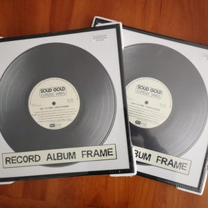 May include: Two framed vinyl records with the text "RECORD ALBUM FRAME" at the bottom. The records are black with white labels that read "SOUD GOLD CLASSIC VINYL". The frames are black with white borders.