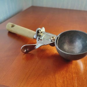 May include: An ice cream scoop with a wooden handle and a metal bowl. The scoop has a lever mechanism for easy release. The handle is light-colored, and the bowl is a darker metal. The scoop is designed for portioning ice cream.