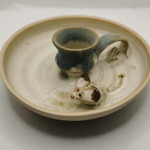 May include: A handcrafted ceramic candle holder with a mouse motif. The holder has a small cup for the candle, a handle, and a decorative mouse figurine. The color scheme includes white, brown, and blue hues.