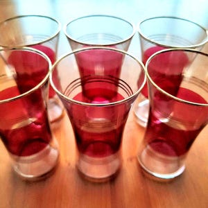 May include: Six vintage red and clear glass cordial glasses with gold rims and decorative bands. The glasses have a flared shape and are arranged on a wooden surface. The red colour is concentrated at the base of the glasses.