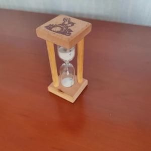 May include: A wooden hourglass with a square top and base, connected by four vertical supports. The top features a carved image of a rabbit. The hourglass contains white sand and is set on a brown surface.