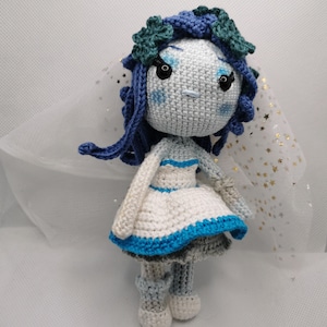 May include: A handmade crochet doll in a bridal theme. The doll has a gray face with blue accents, dark blue hair, and a white dress with blue trim. A white veil with gold stars is attached to the doll's head.