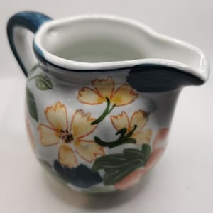 Peut inclure: Un petit pot à lait en céramique avec une base blanche ornée d'un motif floral peint à la main dans des tons de jaune, vert et rose tendre. Le pot possède une anse bleu foncé et un liseré bleu foncé près du bec verseur.