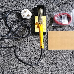 Könnte beinhalten: Ein gelber Lötkolben mit schwarzem Stromkabel und Stecker, ein schwarzer Metallständer mit gelbem Reinigungsschwamm, eine rechteckige braune perforierte Leiterplatte und eine Spule mit rotem Draht in einem durchsichtigen Plastikbeutel.