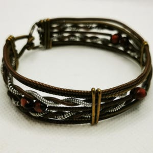 Könnte beinhalten: Ein handgefertigtes Armband aus recycelten Gitarrensaiten. Das Armband hat ein gewebtes Design mit Braun-, Silber- und Bronzetönen. Kleine rote Perlen sind in das Design eingestreut.