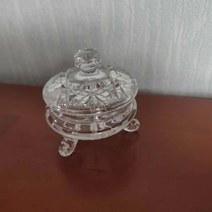 May include: A clear, cut-glass trinket dish with a lid, resting on three small feet. The dish features a detailed, geometric pattern and a knob on the lid for easy handling. The design is classic and elegant, suitable for storing small items.