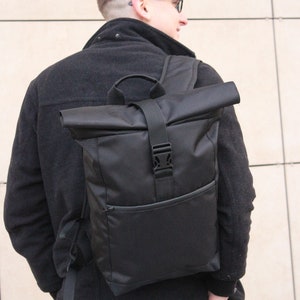 May include: A black roll-top backpack with a buckle closure and a front pocket. The backpack is worn by a person wearing a black jacket and grey pants.