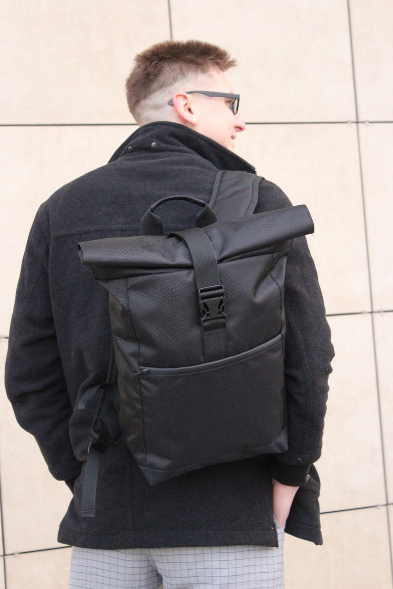 May include: A black roll-top backpack with a buckle closure and a front pocket. The backpack is worn by a person wearing a black jacket and grey pants.