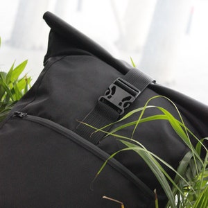 May include: A black roll-top backpack with a zippered pocket and a black buckle closure. The backpack is partially obscured by green foliage.