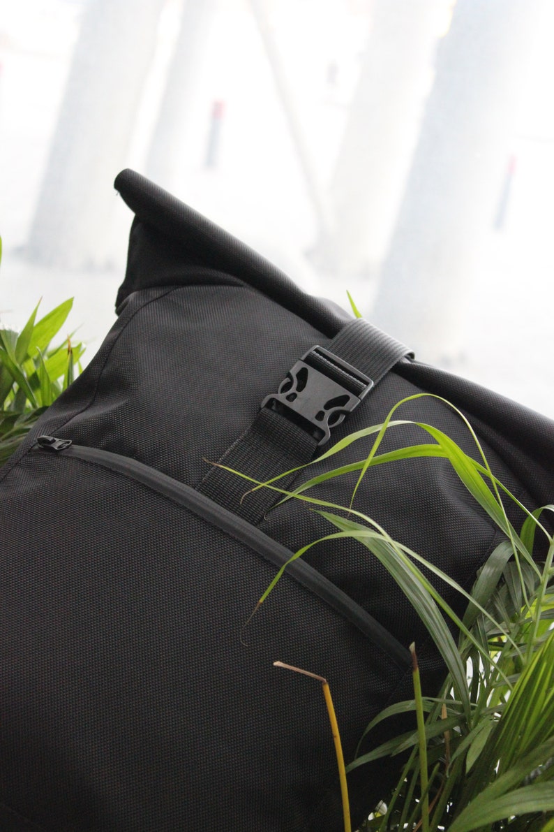 May include: A black roll-top backpack with a zippered pocket and a black buckle closure. The backpack is partially obscured by green foliage.