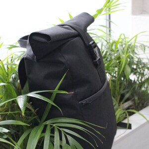 May include: A black roll-top backpack with a zippered pocket and a buckle closure. The backpack is partially obscured by green plants.