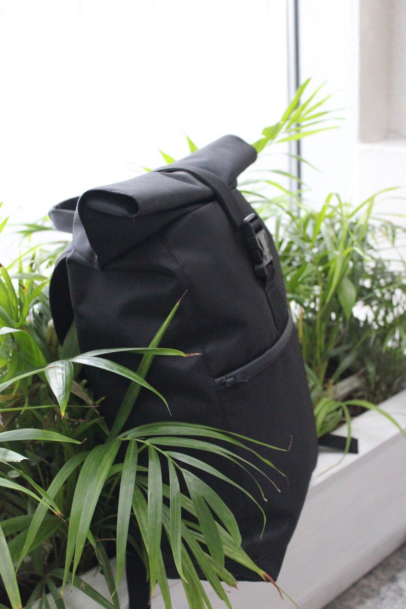 May include: A black roll-top backpack with a zippered pocket and a buckle closure. The backpack is partially obscured by green plants.