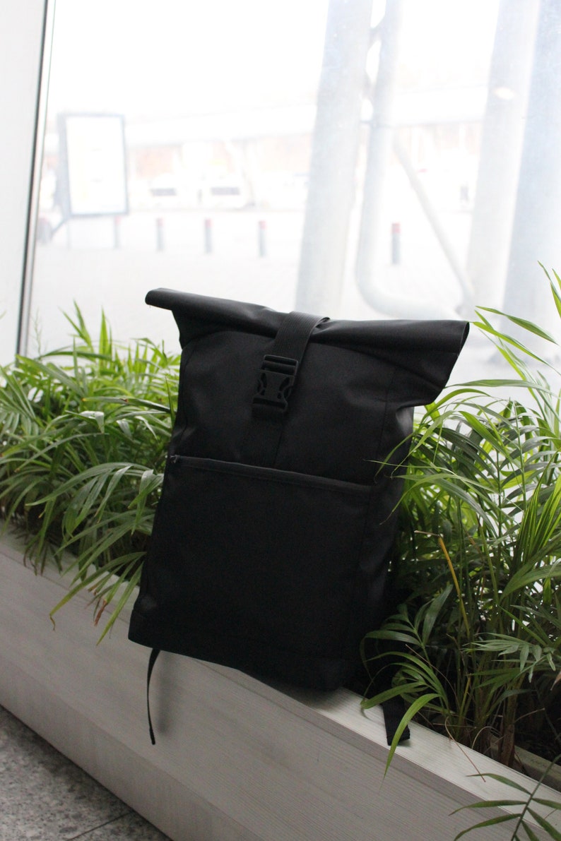 May include: A black roll-top backpack with a buckle closure. The backpack is sitting on a white surface in front of a window.