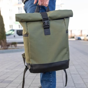 May include: Olive green roll-top backpack with black accents. The backpack features a black buckle closure, a black leather bottom, and adjustable straps. The bag is held by a person in blue jeans.