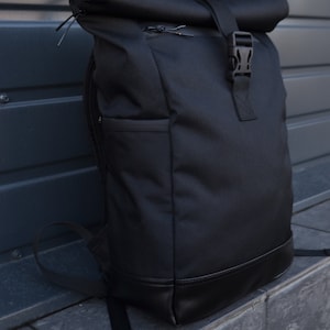 Op de afbeelding: Een zwarte roll-top rugzak met een gespsluiting en een leren rand. De rugzak is gemaakt van een duurzaam, waterbestendig materiaal.