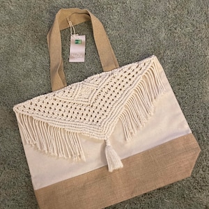 Peut inclure: Un sac fourre-tout en toile blanche avec un dessus en crochet orné d'une frange et d'un pompon. Le sac a un fond en jute brun et des anses en jute brun.