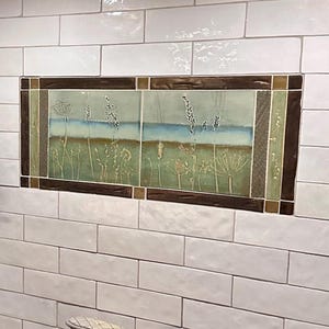 Puede incluir: Una tetera de cobre sobre una estufa de gas negra con el logo de Samsung. La pared tiene azulejos blancos tipo metro, un estante de madera oscura, un grifo negro montado en la pared y un mural decorativo de azulejos con una escena de prado.