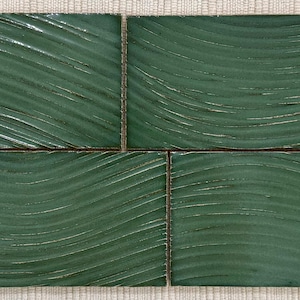 Puede incluir: Cuatro baldosas rectangulares de cerámica verde oscuro con un diseño ondulado y texturizado. Las baldosas están dispuestas en un cuadrado, con líneas de lechada visibles. Las baldosas están sobre una superficie tejida beige claro.