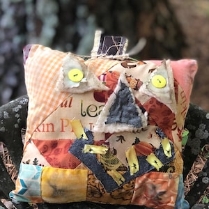 May include: A patchwork pillow with a pumpkin design. The pillow features various fabric patterns, including orange gingham, floral prints, and denim. Yellow buttons and ribbon accents add detail. The pillow is designed to look like a pumpkin.