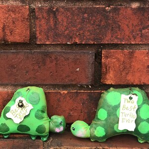 May include: Two handmade green turtle plush toys with painted green spots. Each turtle has a tag that reads "Best Turtles Ever". The turtles are displayed against a red brick wall.