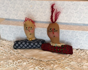 May include: Two handmade fabric dolls with painted faces and yarn hair. One doll wears a navy blue floral print dress, the other a red patterned dress. Both dolls have a decorative collar and are displayed on a lace doily.