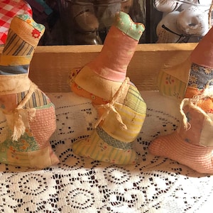 May include: Three handmade fabric boot ornaments in a wooden tray. The boots are made of patchwork fabric in various colors and patterns, including pink, yellow, blue, and green. Each boot is tied with a string and has a small, curved top.