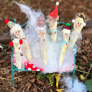 May include: A whimsical display of six snowman figures, each crafted from a light-colored material and adorned with festive hats, scarves, and painted faces. They are arranged on a miniature teal bench, surrounded by white netting and a red and white polka-dot fabric.
