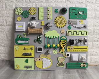 Puede incluir: Un tablero de actividades de madera para niños con diferentes texturas, colores y formas. Cuenta con un cocodrilo verde, una abeja amarilla, un zapato verde, una nube verde con cuerdas y bolas, un laberinto verde, un sol amarillo, un interruptor de luz blanco, una toma de corriente blanca, una cadena, una cerradura, una campana, un spinner y los números del 1 al 9.