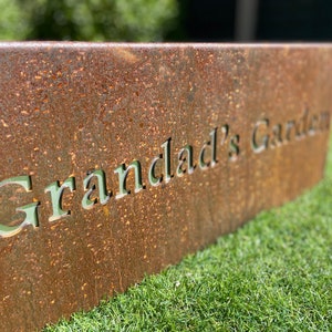 May include: A rusty metal sign with the words "Grandlad's Garden" cut out in the metal. The sign is sitting on a bed of green grass.